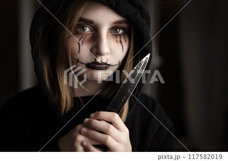 Spooky little girl holding knife and looking at blade. Scary child kid in black and Halloween dark makeup 117582019