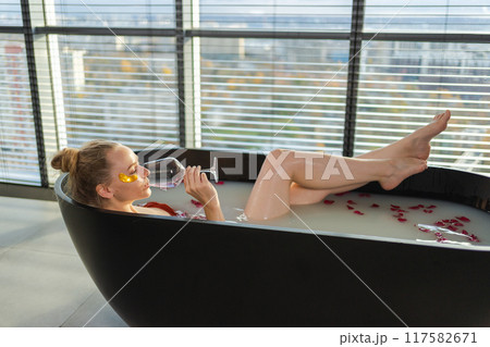 Spa relaxation. Woman lying in bath with holding glass with red wine. Girl relaxing in bathroom at home. Pretty female taking hot bath drinking wine. Stress relief. Rest after hard working day 117582671