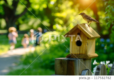 Wooden birdhouse on a fence with a bird perched on top, set against a sunny park scene with children 117582782