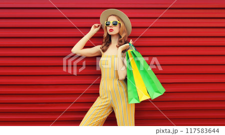 Shopping day, stylish beautiful young woman with shopping bags in summer hat on red wall background 117583644