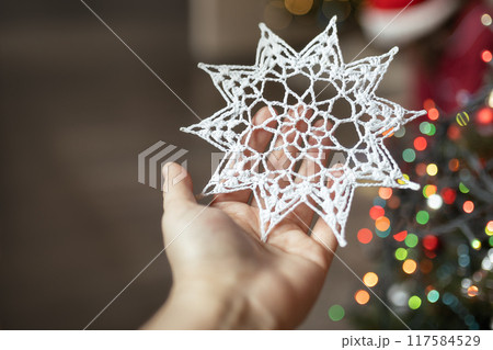 knitted white handmade snowflake with ornaments in hand on background of Christmas tree at home 117584529