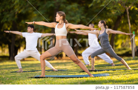 Girl performing Warrior pose during group yoga session in park Girl performing Warrior pose during group yoga session in park 117587858