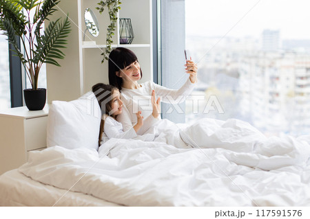 Mom and daughter lying in bed having video call on phone Mom and daughter lying in bed having video call on phone 117591576