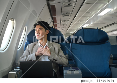 Young businessman looking out the window with hand resting on his chest, experiencing aerophobia or fear of flying Young businessman looking out the window with hand resting on his chest, experiencing aerophobia or fear of flying 117591914
