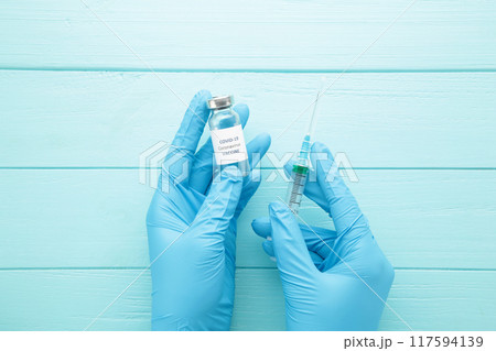 Bottle with vaccine against covid 19 and syringe in hand of researcher wearing gloves on blue background. Bottle with vaccine against covid 19 and syringe in hand of researcher wearing gloves on blue background. 117594139
