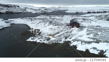 Lonely building on Antarctica rocky coast, snow covered island bay, mountains in background. Aerial view of isolated polar station house surrounded by snowy terrain. Travel expedition to South Pole Lonely building on Antarctica rocky coast, snow covered island bay, mountains in background. Aerial view of isolated polar station house surrounded by snowy terrain. Travel expedition to South Pole 117594391