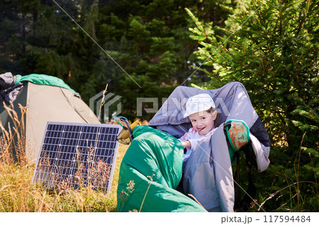 Young child playing near tourist tent with photovoltaic solar panel in summer. On background of dense forest. Integration of renewable energy in outdoor camping activities. Young child playing near tourist tent with photovoltaic solar panel in summer. On background of dense forest. Integration of renewable energy in outdoor camping activities. 117594484