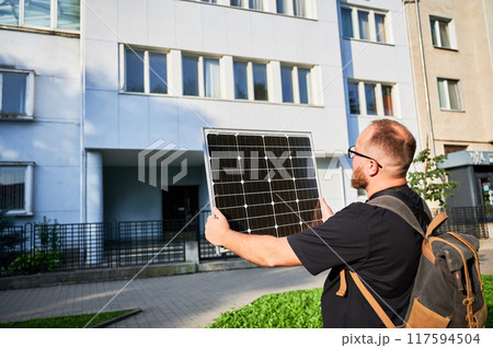 Man holding photovoltaic solar panel in front of historical building. Concept of integration of sustainable renewable energy sources into architecture. Man holding photovoltaic solar panel in front of historical building. Concept of integration of sustainable renewable energy sources into architecture. 117594504