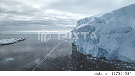 Massive glacier in Antarctica peacefully floats polar ocean. Towering snow covered iceberg rises above cold, dark water. Ice wall formation under moody sky. Antarctica winter landscape panorama Massive glacier in Antarctica peacefully floats polar ocean. Towering snow covered iceberg rises above cold, dark water. Ice wall formation under moody sky. Antarctica winter landscape panorama 117594534