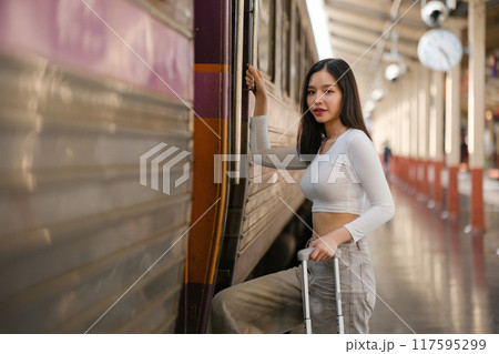A woman is standing in front of a train with a suitcase 117595299