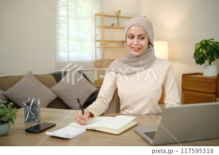 A woman wearing a head scarf is sitting at a desk with a laptop and a notebook 117595318