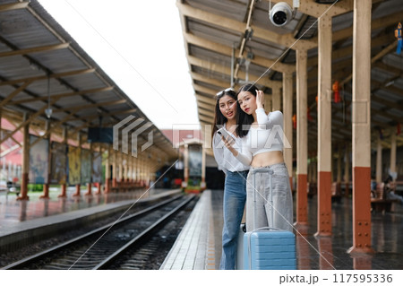 Two women standing at a train station with their luggage 117595336