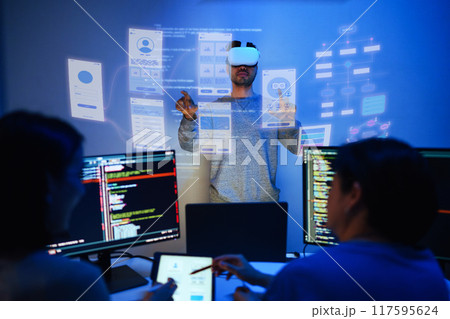 developer uses a VR headset to interact with projected interface designs, while colleagues discuss and code on screens at office 117595624