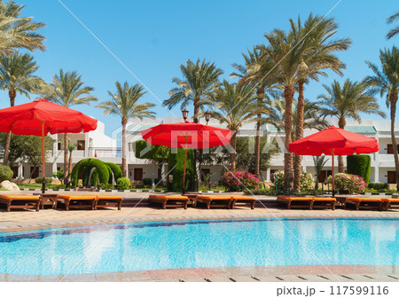 Outdoors swimming pool with bright red umbrellas near hotels or residential houses and palms. Outdoors swimming pool with bright red umbrellas near hotels or residential houses and palms. 117599116