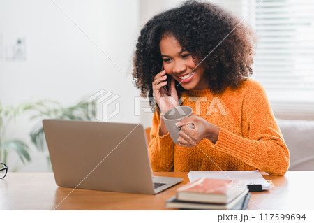 Smiling African American woman enjoys phone call while holding coffee cup and working on laptop at home. 117599694