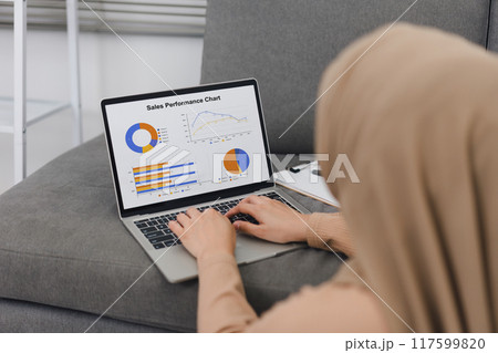 Muslim woman wearing hijab uses laptop to analyze sales performance charts while sitting on couch. 117599820