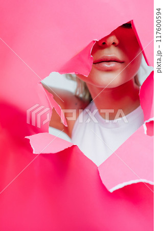 View of beautiful young woman with plumped lips through hole in pink paper. View of beautiful young woman with plumped lips through hole in pink paper. 117600594