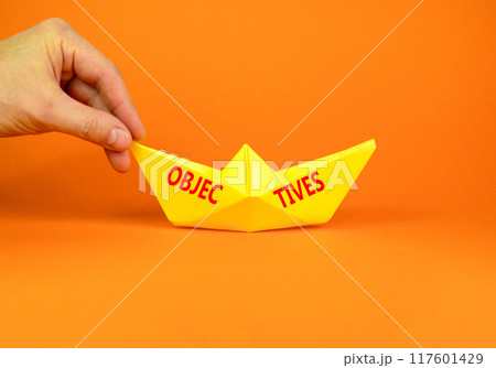 Objectives symbol. Concept word Objectives on beautiful yellow paper boat. Beautiful orange background. Businessman hand. Business objectives concept. Copy space. 117601429