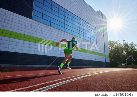 Focused and fast, athlete in green races along track, with the sleek building facade adding modern touch to his sprint. 117601548