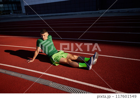 After tough run, athlete sits on the track, deep in thought, his green outfit contrasting sharply with vibrant red surface beneath him. 117601549