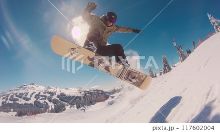 Snowboarder in sportswear performing dynamic jump, kicking up spray of snow against backdrop of clear blue skies and snowy mountain peaks 117602004