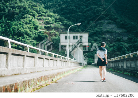 橋の上の道路を歩いてくセーラー服の女の子の後ろ姿 117602200