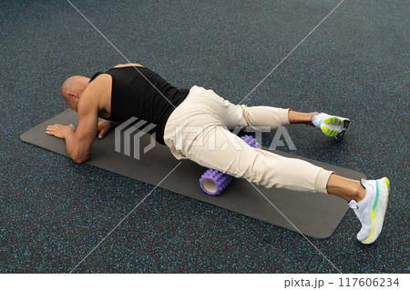 Athlete man exercising and stretching legs on foam roller on sports ground. Outdoor activity. Morning routine. Public park. Athlete man exercising and stretching legs on foam roller on sports ground. Outdoor activity. Morning routine. Public park. 117606234
