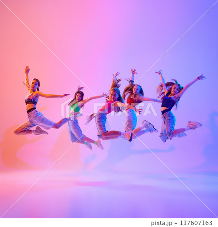 Dynamic photo of young girls, teenagers performing, jumps in mid-air in neon light against gradient pink background. 117607163