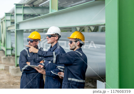 Close up side view group of engineer or factory technician worker team stand near petrochemical gas pipeline and use tablet to discuss about their work with one point forward. Close up side view group of engineer or factory technician worker team stand near petrochemical gas pipeline and use tablet to discuss about their work with one point forward. 117608359