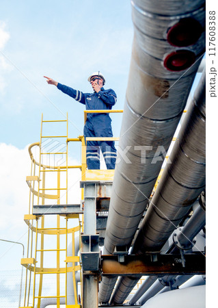 Vertical image of factory engineer or technician worker man use walkie-talkie to contact co-worker also point forward with stand on high state over pipeline of petrochemical gas in workplace. 117608388