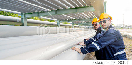 Side view of two engineer or factory technician workers stand beside petrochemical gas pipeline also touch and look at camera with warm light. 117608391