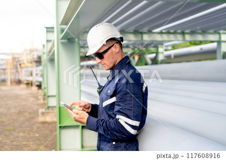 Side view of engineer or technician worker leaning against the petrochemical pipeline and enjoy with his mobile phone. Side view of engineer or technician worker leaning against the petrochemical pipeline and enjoy with his mobile phone. 117608916