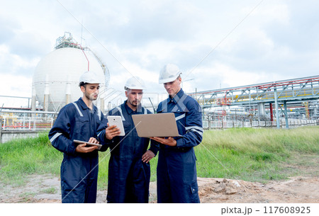 Group of engineer or technician workers stand and look to laptop in front of petrochemical factory with cloudy sky. Group of engineer or technician workers stand and look to laptop in front of petrochemical factory with cloudy sky. 117608925
