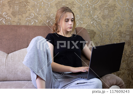 Young woman is engaged in remote work on her laptop while comfortably seated in a cozy living room space Young woman is engaged in remote work on her laptop while comfortably seated in a cozy living room space 117609243