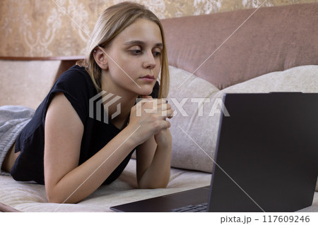 Girl student lies on sofa on her stomach and uses laptop while listening to webinar, distance modern learning of the future generation Girl student lies on sofa on her stomach and uses laptop while listening to webinar, distance modern learning of the future generation 117609246