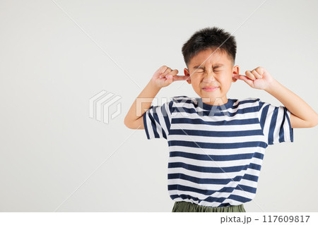 Asian young kid boy expression of pain and frustration is evident as she covers ears and closes her eyes with fingers to escape the loud noise in studio shot isolated white background, primary child 117609817