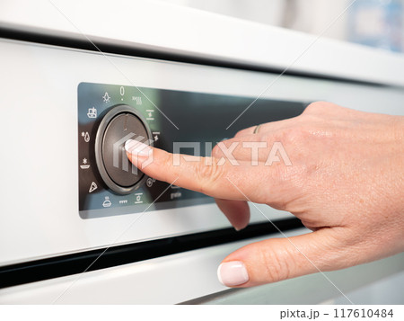 Woman adjusting modern white oven in kitchen.  Female hand pushing and turning knobs on oven control panel 117610484