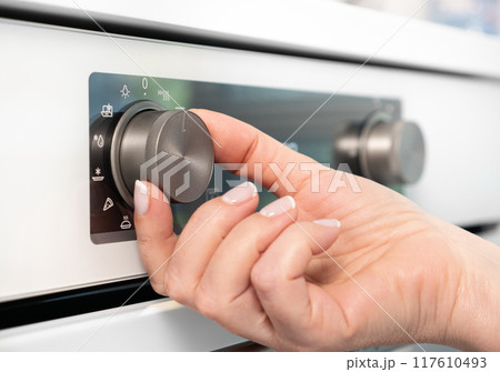 Woman adjusting modern white oven in kitchen. Female hand pushing and turning knobs on oven control panel Woman adjusting modern white oven in kitchen. Female hand pushing and turning knobs on oven control panel 117610493