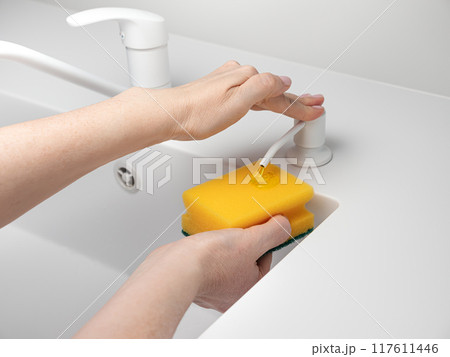 Female hand press on built-in dispenser in the sink with detergent. Woman washing dishes by yellow sponge with soap in white kitchen Female hand press on built-in dispenser in the sink with detergent. Woman washing dishes by yellow sponge with soap in white kitchen 117611446