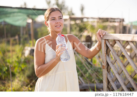 Girl stands near garden bed and drinks water from bottle 117618447