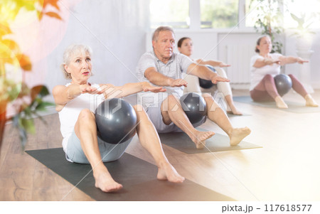 Mature people with young coach sit on karemat, pulled leg to themselves and clamped softball in knee 117618577