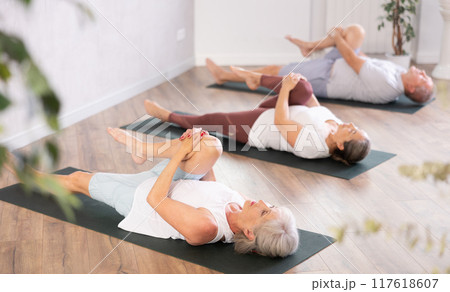 Class of mature women stretching gluteal muscle while lying on back during yoga Pilates workout on mats in gym studio 117618607