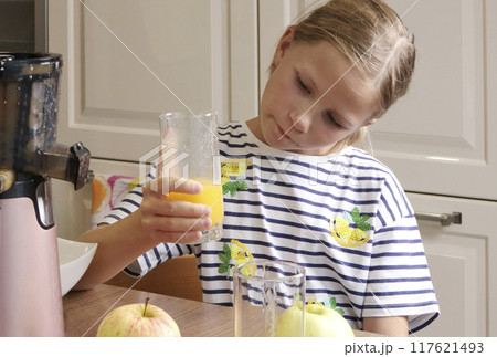 Little girl look at glass of orange juice. Electric juicer on table in kitchen at home. Food and health. Little girl look at glass of orange juice. Electric juicer on table in kitchen at home. Food and health. 117621493