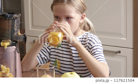 Young girl drink orange juice from glass in kitchen at home. Electric juicer on the table. Nutrition and health. Young girl drink orange juice from glass in kitchen at home. Electric juicer on the table. Nutrition and health. 117621504