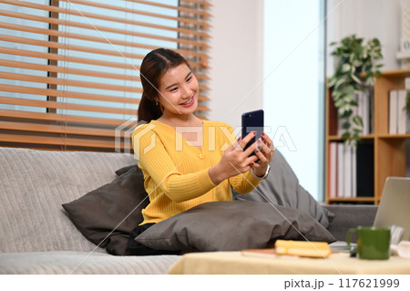 Happy young woman enjoying chatting with her friends using smartphone on sofa at home Happy young woman enjoying chatting with her friends using smartphone on sofa at home 117621999