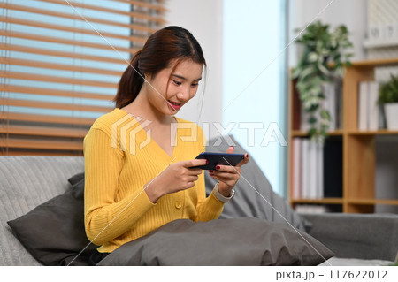 Young pretty asian woman relaxing on couch at home and playing video games on her smartphone 117622012