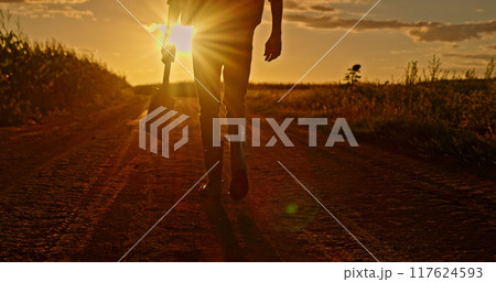 Farmer's legs in boots walking through a field at sunset, carrying a shovel. 117624593