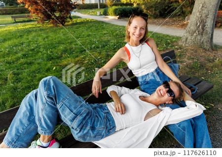Pair smiling happy young adult women enjoy sunny day together sit relaxing on bench in city park outdoors. Female persons wearing casual denim white tops portrait. Best friends friendship outside 117626110