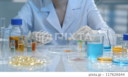 A scientist is pouring an orange solution into a petri dish for testing. Surrounding them are flasks containing test samples and substances. Creative pharmacy or health care concept. A scientist is pouring an orange solution into a petri dish for testing. Surrounding them are flasks containing test samples and substances. Creative pharmacy or health care concept. 117626844