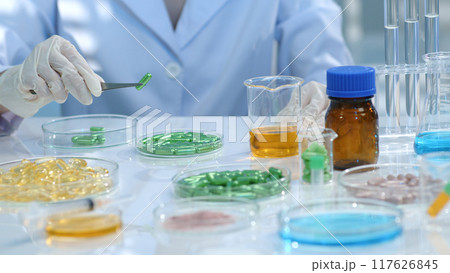 on the research table, many drug samples are stored in petri dishes. The researcher is using tweezers to pick up a green pill and examine it. Research landscape for medicinal products 117626845